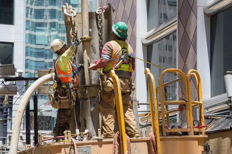 Two construction workers in safety vests lower a steel I-beam into an excavated shaft on an urban street.