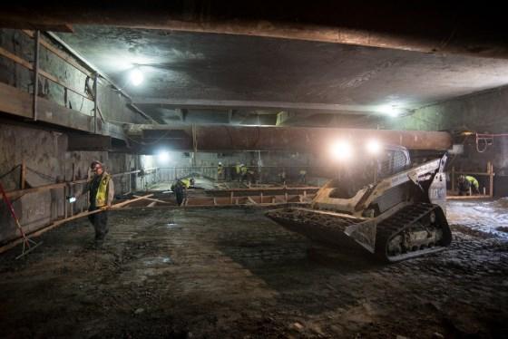 Workers grade soil inside underground transit station box during construction.