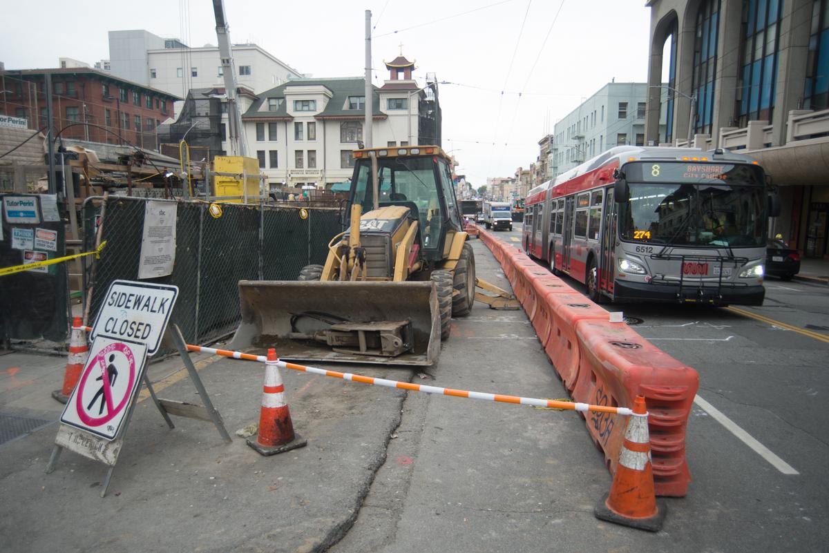 Construction equipment and barriers block a city sidewalk next to a transit bus on a street.
