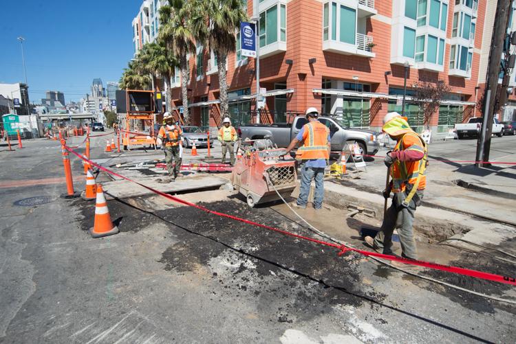 Construction workers in safety gear excavate a street intersection with orange cones and red barriers marking the work zone in San Francisco.