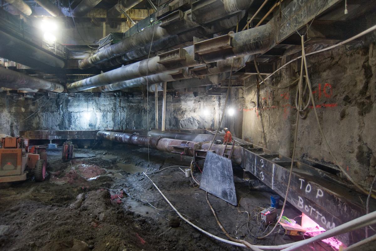 Underground station construction with exposed pipes, steel reinforcement, and excavation equipment in concrete cavern.