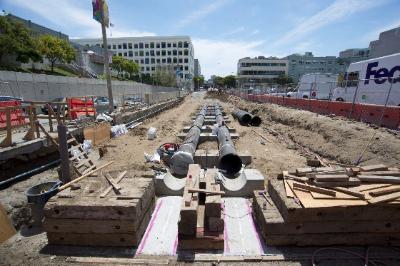 Construction site showing excavated tunnel section with utility pipes and roof deck installation in urban area.