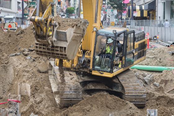 Yellow excavator with bucket raised, moving soil and debris at urban construction site.
