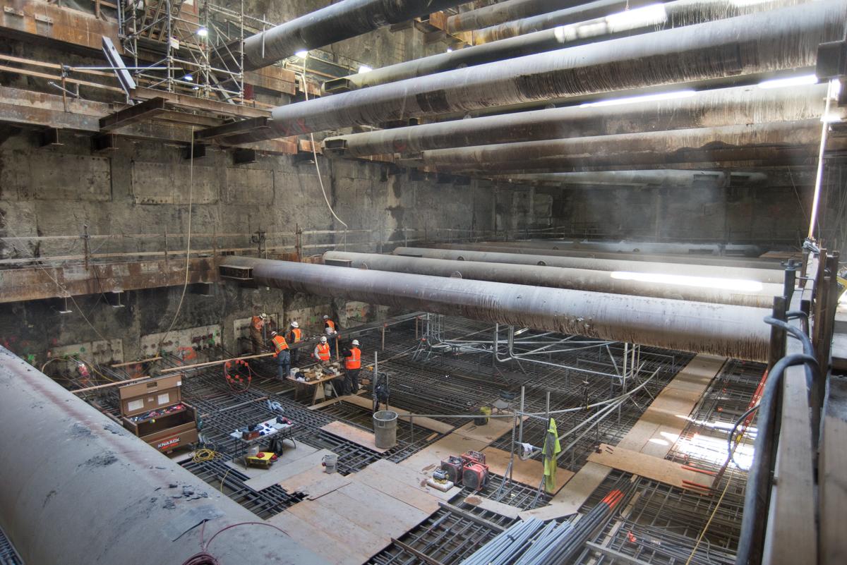 Workers in safety gear install utility systems in Chinatown Station underground construction pit with large pipes overhead.