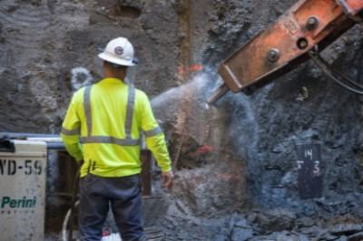 Worker in safety gear spraying water on concrete wall during excavation work to control dust.