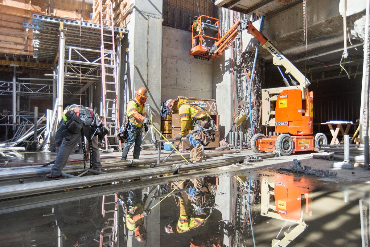 Construction workers assemble scaffolding inside Yerba Buena/Moscone Station headhouse with aerial lifts and equipment.