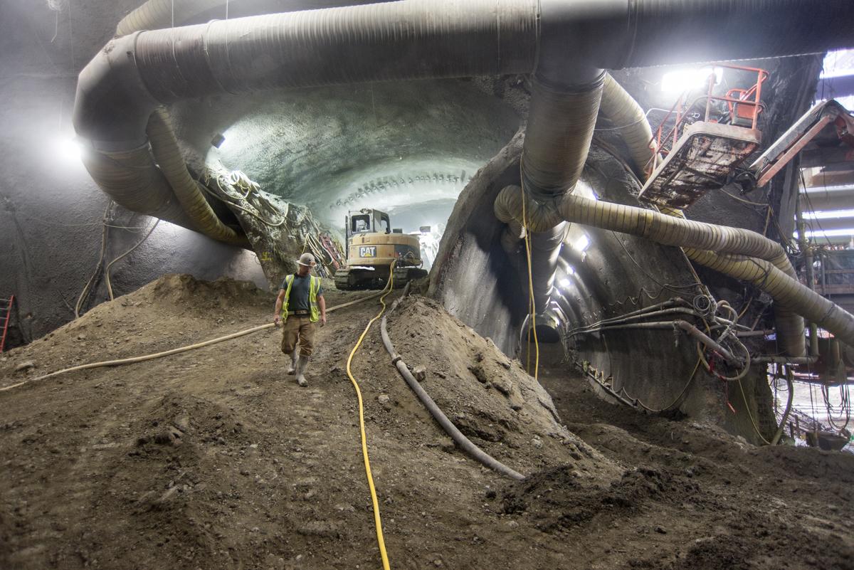 Workers excavate tunnel ceiling in underground transit construction site with heavy equipment and lighting.