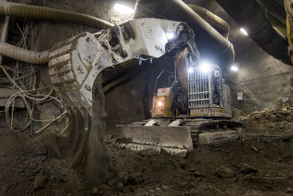 Excavator operating underground in tunnel, moving soil at night with bright work lights illuminating the concrete cavern.