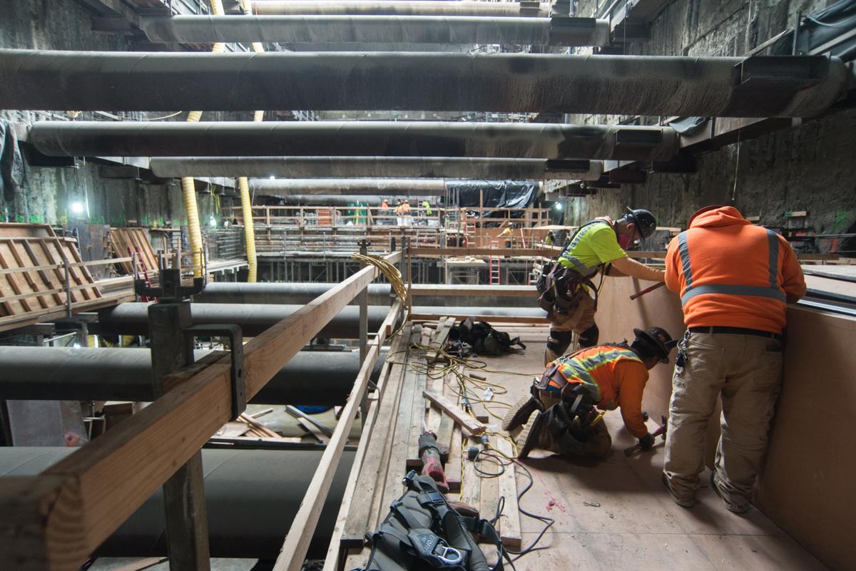 Workers in safety vests perform construction work on an underground transit shaft with exposed pipes and structural beams.