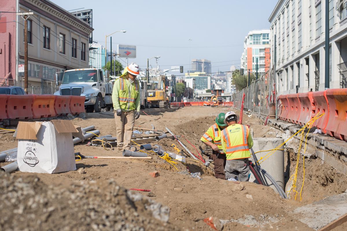 Construction workers in safety vests excavate trenches on an urban street lined with red barriers and equipment.