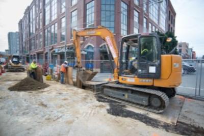 Yellow excavator digging trench for sewer line installation on city street with construction workers and brick building in background.