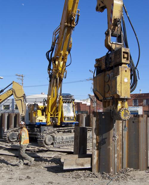 Heavy construction equipment including yellow excavators and pile driving machinery at a Central Subway construction site.