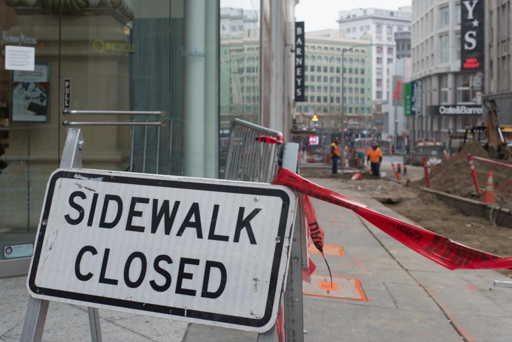 Sidewalk Closed sign with red and white barrier tape blocking pedestrian access on urban street.