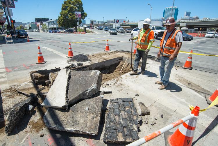 Workers in safety vests examine exposed cobblestones and underground utilities at a street construction site.