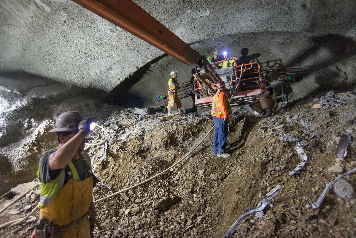 Workers in safety gear apply shotcrete inside a tunnel cavern during construction, with equipment and steel supports visible.