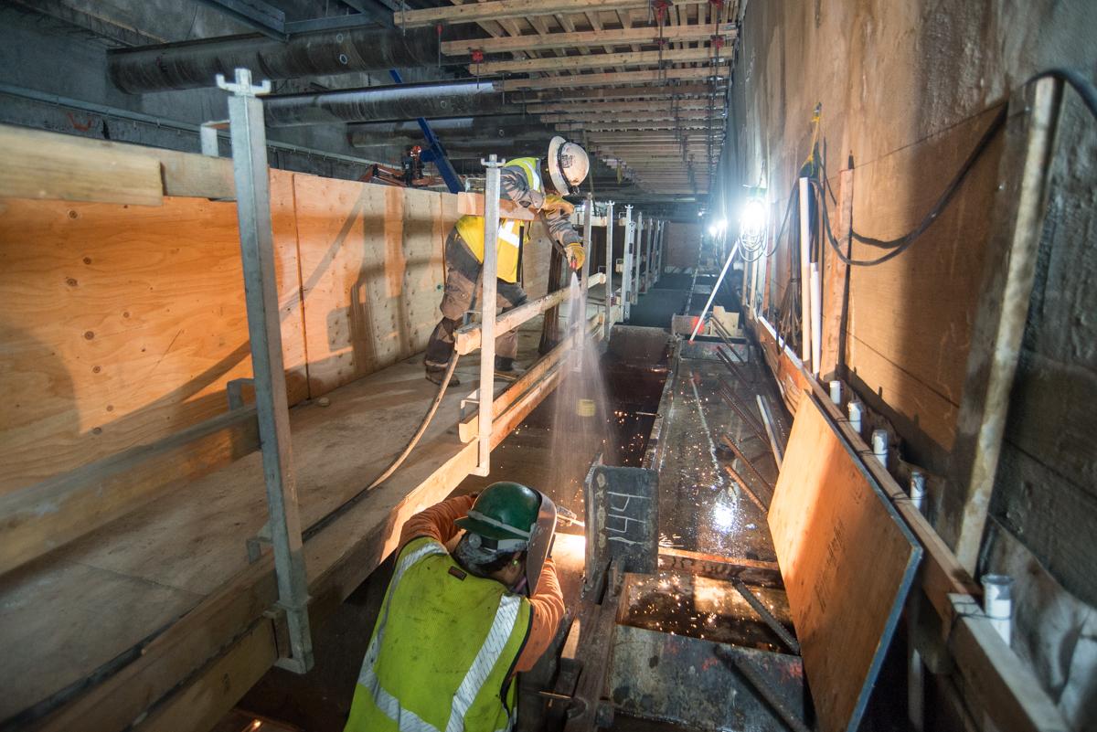 Workers in safety gear performing welding and steel installation inside an underground transit station excavation.