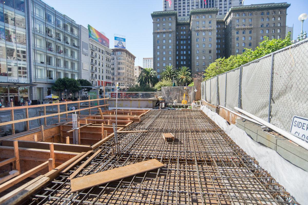 Construction site with rebar and concrete forms at underground station entrance, surrounded by safety fencing and wooden bracing.