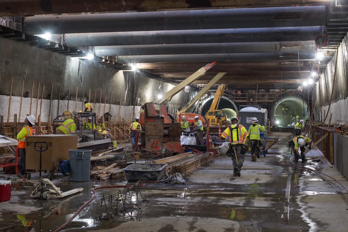 Workers in safety vests construct concrete forms and steel bracing inside an underground transit station tunnel.