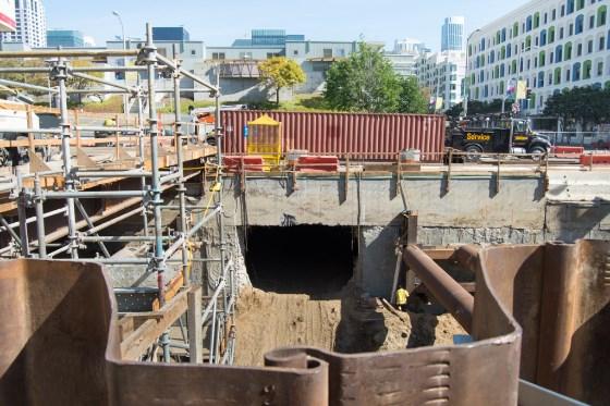 Excavation pit beneath 4th Street showing interior station construction with metal barriers and equipment surrounding the work area.