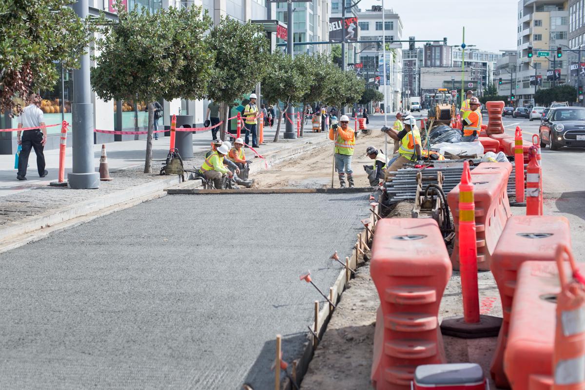 Construction workers in safety vests install steel sheet piles along a city street with traffic barriers and equipment visible.