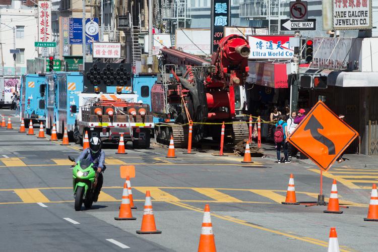 Street construction site with drill rig, utility vehicles, orange traffic cones, and green motorcycle on closed urban street.