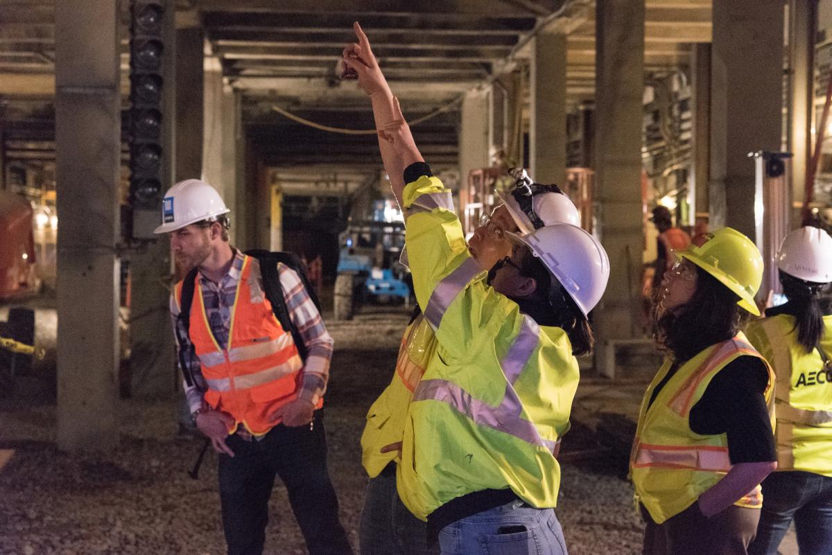 Construction workers in safety vests and hard hats inspect structural work inside a transit tunnel with concrete pillars and overhead infrastructure.