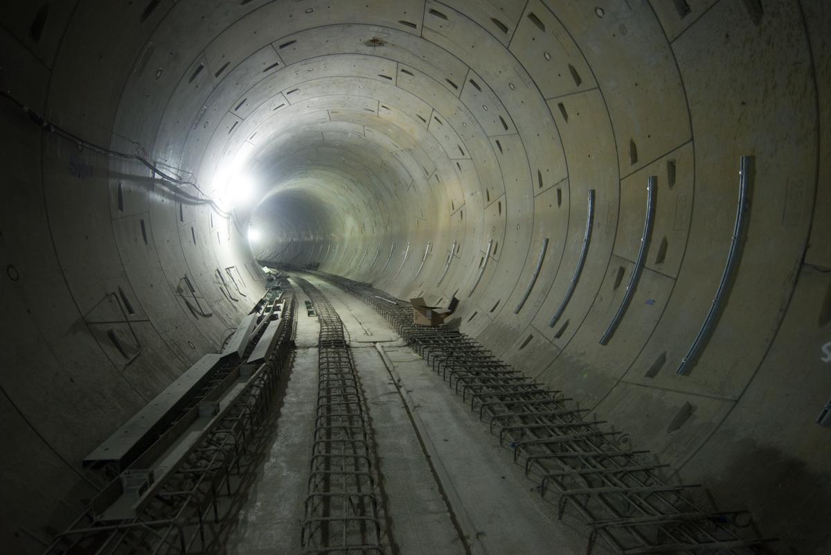 Interior view of subway tunnel under construction with steel reinforcement and utility conduits along walls.