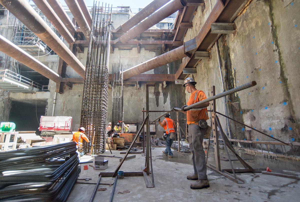Construction workers in orange vests assemble rebar cages and concrete forms inside an underground station excavation.