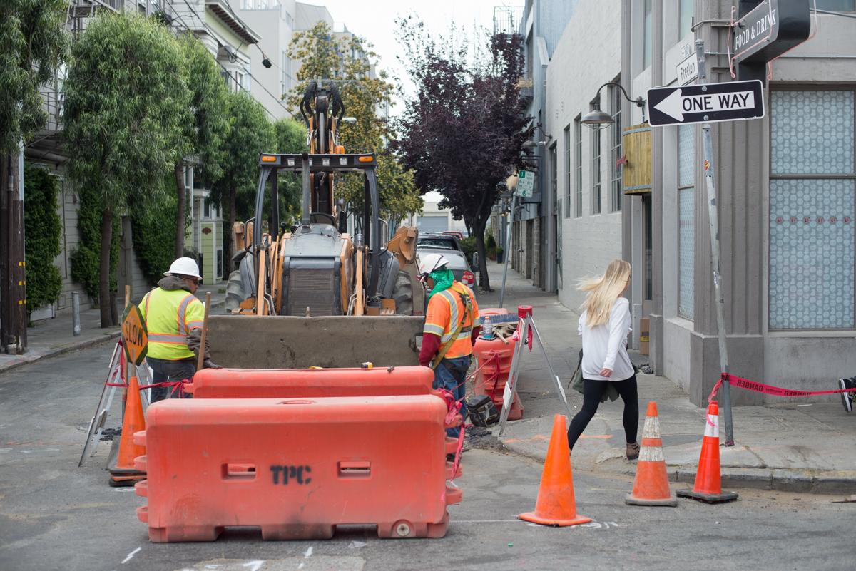 Construction crew conducting utility trenching work on a city street with heavy equipment and safety barriers.