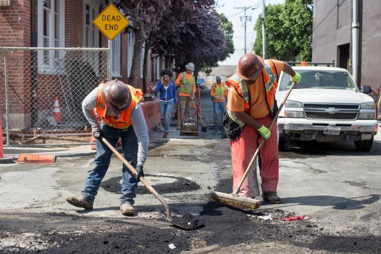 Workers applying asphalt to street surface on 4th Street, with construction crew and vehicles in background.
