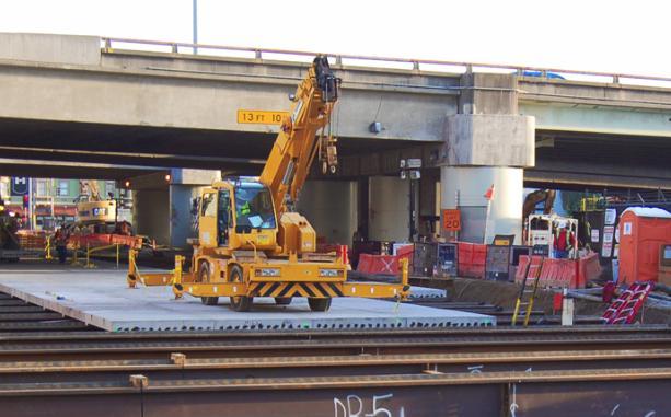 Yellow crane on flatbed truck installing deck panels over tunnel launch box on 4th Street.
