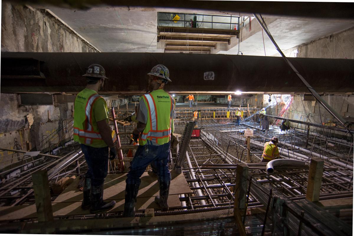 Construction workers in safety vests inspect reinforced steel rebar framework at underground transit station.