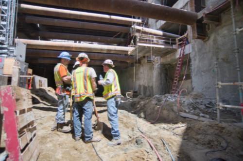 Three construction workers in hard hats and safety vests stand in an underground excavation site with steel support beams overhead.