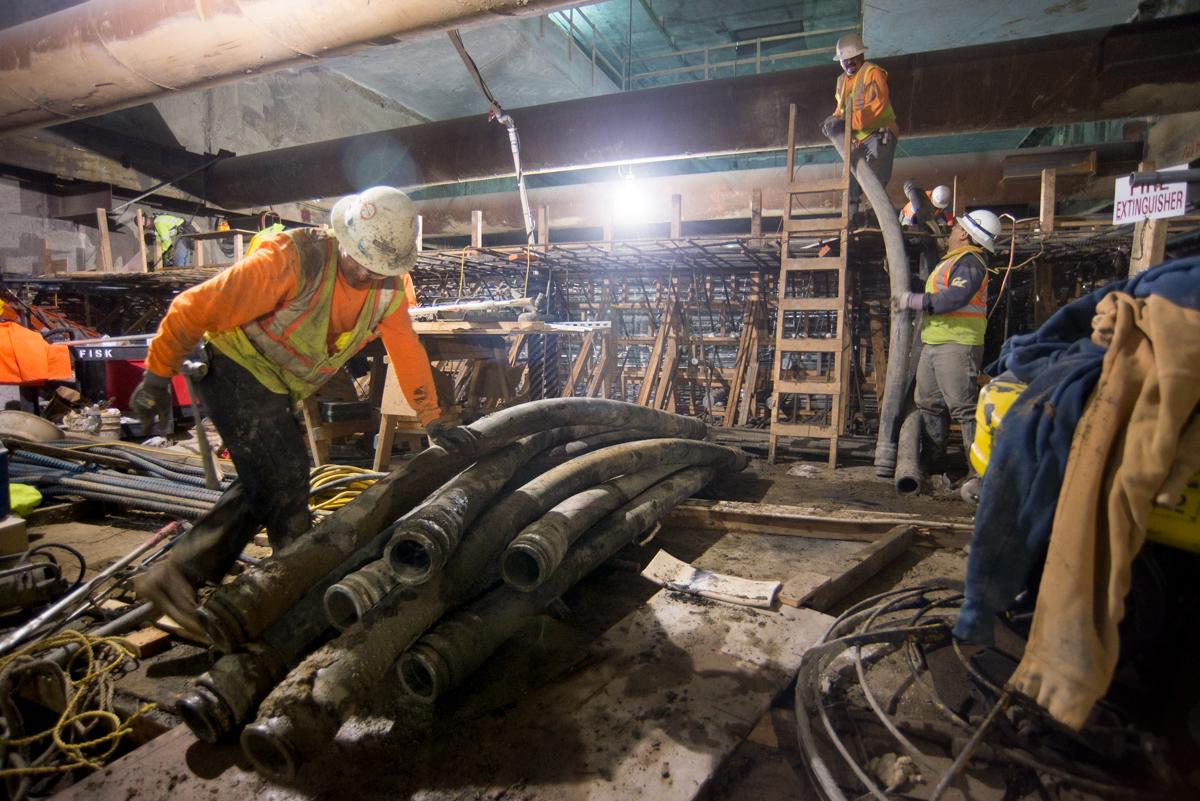 Workers stack concrete pump hose sections at underground transit construction site.