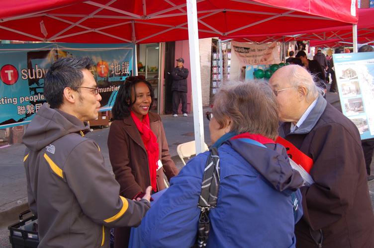 Four people conversing under red tent canopy at outdoor community event with transit signage visible.