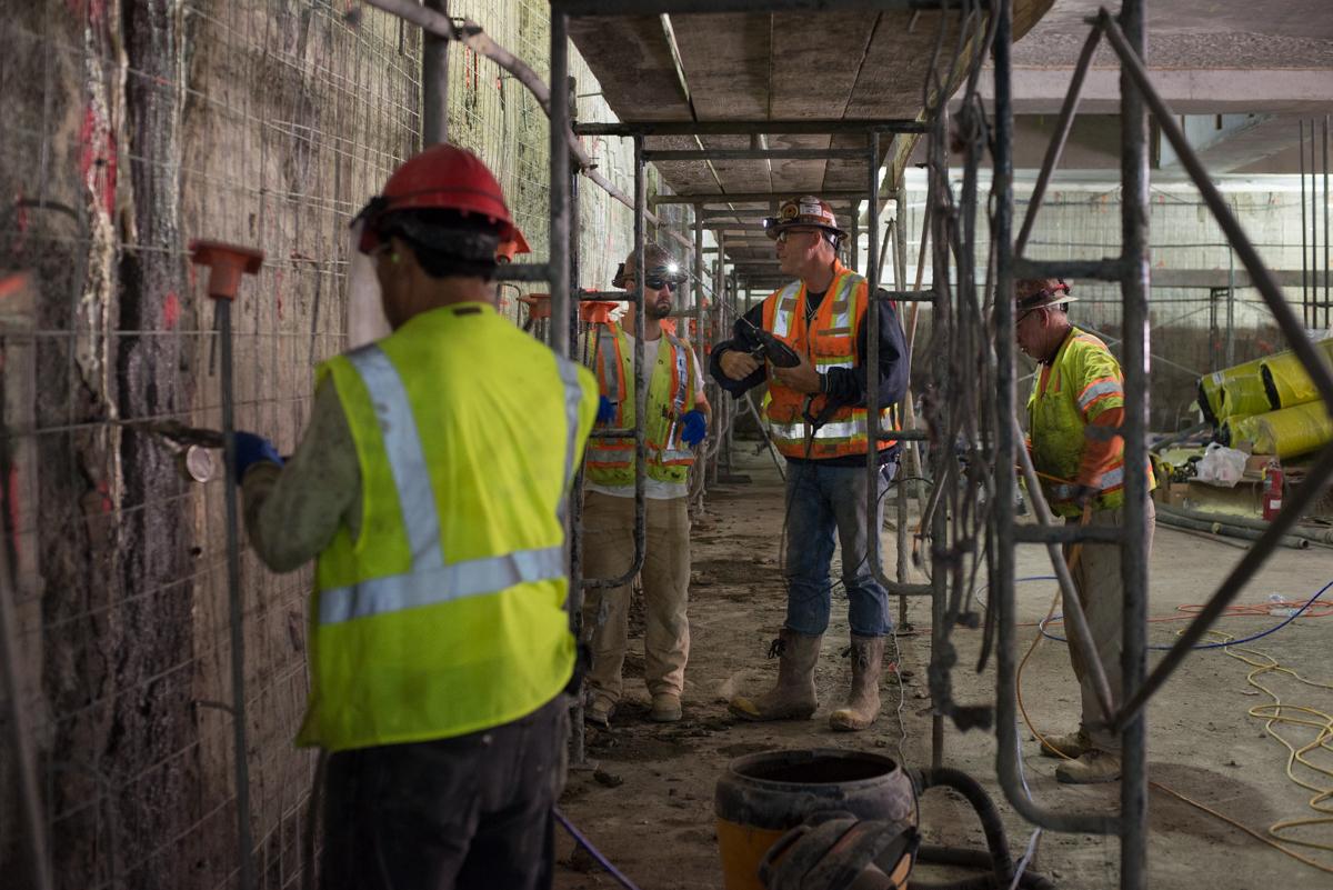 Construction workers in safety vests and hard hats conduct work inside a transit station under temporary metal scaffolding.