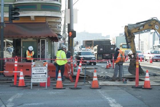 Construction workers in safety vests manage street closure at urban transit construction site with equipment and traffic cones.