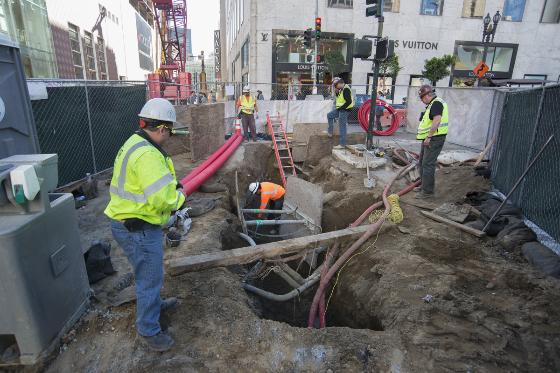 Construction workers in safety gear uncover utility lines in an urban excavation pit.