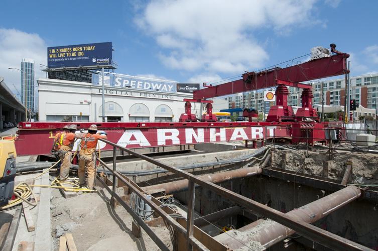 Red gantry crane positioned over 4th Street at Harrison Street for Central Subway construction, with workers and equipment visible below.