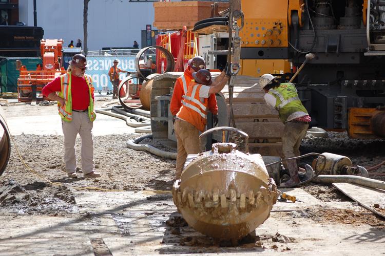 Workers in safety vests operate equipment at a construction site with large cylindrical pipes and heavy machinery.