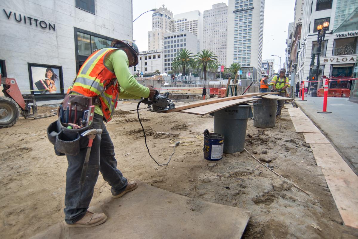 Construction worker in safety vest operating power tool at underground transit station excavation site on urban street.
