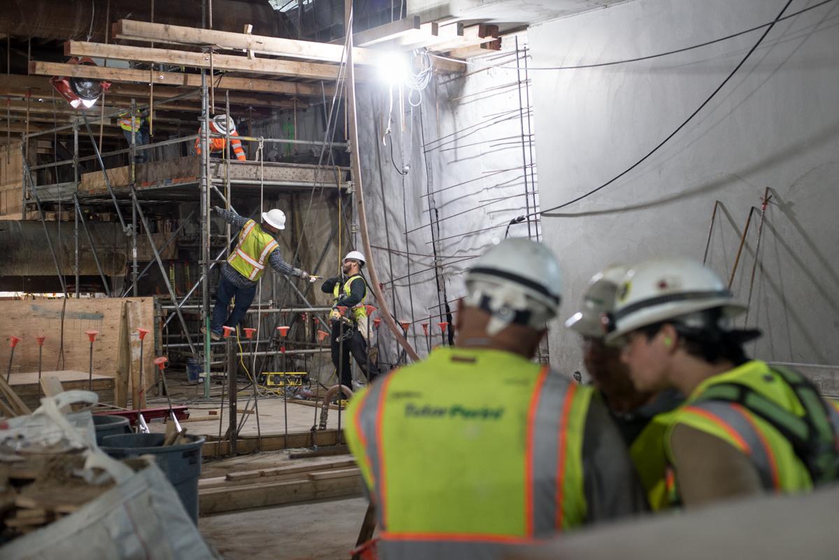 Construction workers in safety vests and hard hats assemble scaffolding on a building mezzanine level.