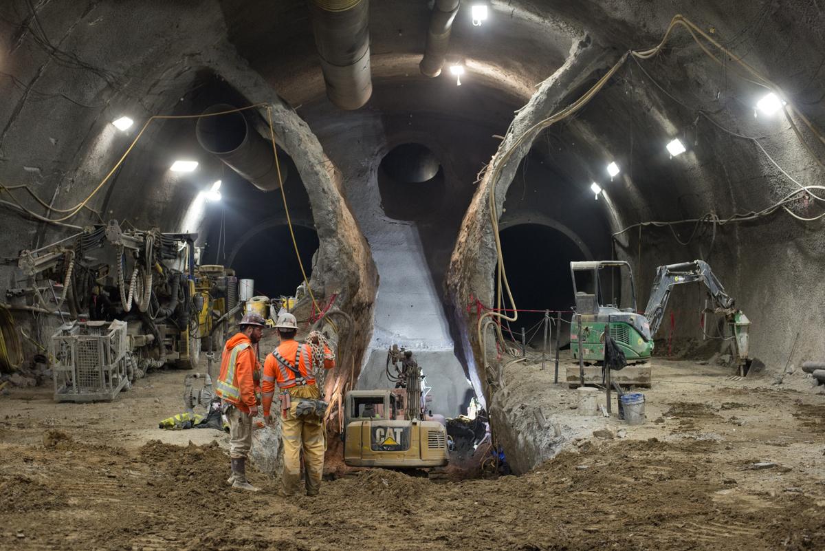 Workers in orange vests stand near a CAT bulldozer inside an underground tunnel excavation site with twin subway tunnels visible ahead.
