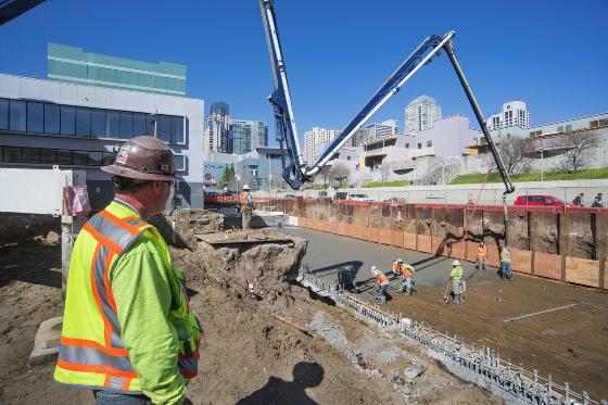 Construction worker in safety gear observing concrete pump operation at urban construction site with city skyline.