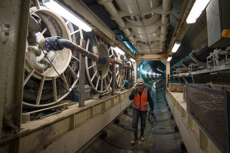 Worker in orange safety vest carries equipment through tunnel past large circular tunnel boring machine components.