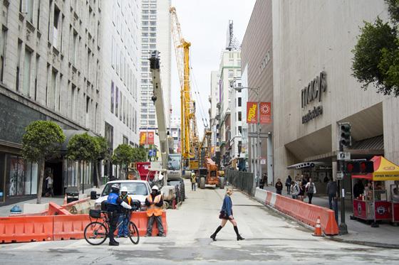 Construction site on Stockton Street with orange barriers, crane, and pedestrians walking past closed lanes.