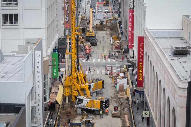 Aerial view of construction equipment and workers on Stockton Street between tall buildings, with yellow drilling rigs and safety barriers visible.
