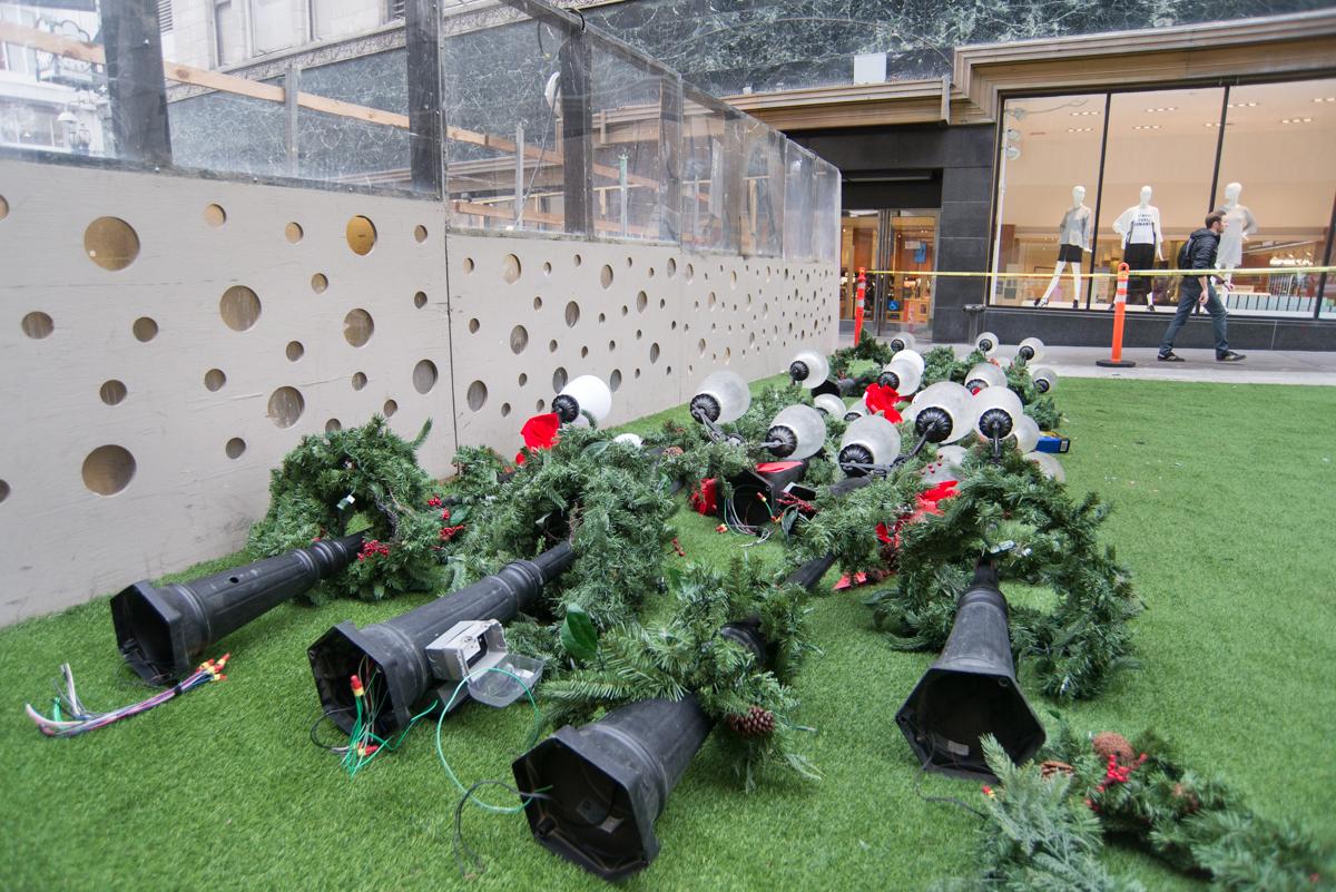 Decorative green wreaths with red bows and vintage-style lamp posts arranged on grass near a perforated metal wall and storefront.