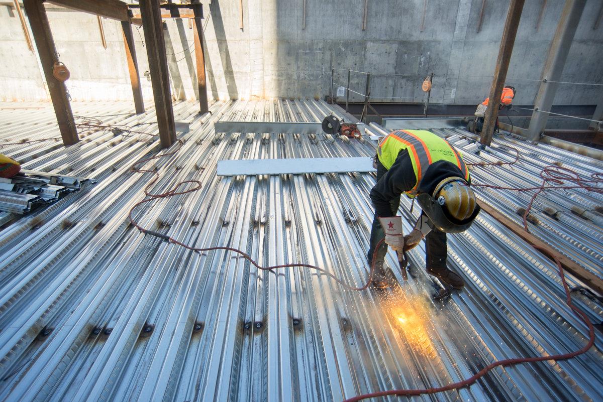 Worker in safety gear welding corrugated steel floor panels inside Central Subway tunnel construction site.