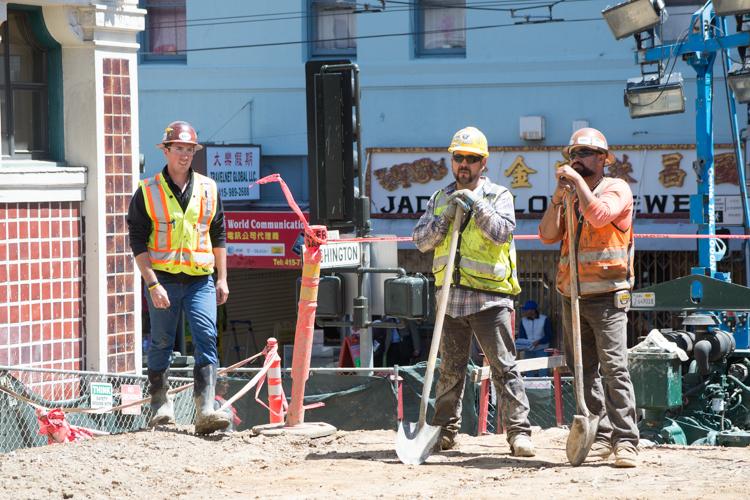 Construction workers in hard hats and safety vests stand at a Chinatown Station construction site with equipment and delivery truck.